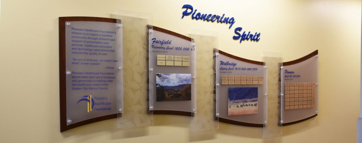 A wall display titled "Pioneering Spirit" features four panels with text, images, and charts. The first panel has text and a logo, the second and third include images and charts, and the fourth has a chart. The background wall is painted a light beige color.