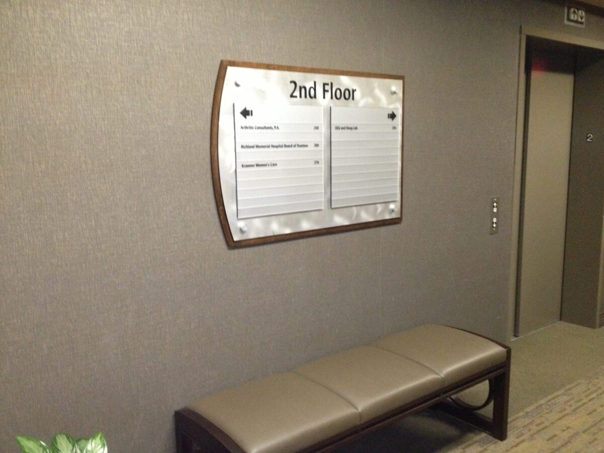 A hallway with a directory sign mounted on a grey wall indicating "2nd Floor." The sign lists various room numbers and details. Below the sign is a brown bench with a cushioned seat. To the right, there is an elevator door and a control panel.