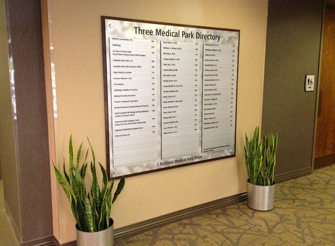 A large medical directory sign titled "Three Medical Park Directory" is mounted on a beige wall. It lists various offices and room numbers. Two potted plants with long, narrow leaves are placed on either side of the sign on the patterned carpet floor.