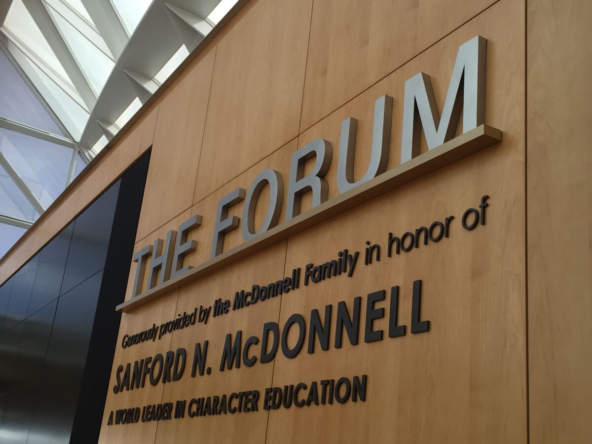 A wooden wall with mounted metal letters reading: "THE FORUM." The wall also displays the text: "Generously provided by the McDonnell Family in honor of Sanford N. McDonnell, A World Leader in Character Education." Brightly lit ceiling visible in the background.