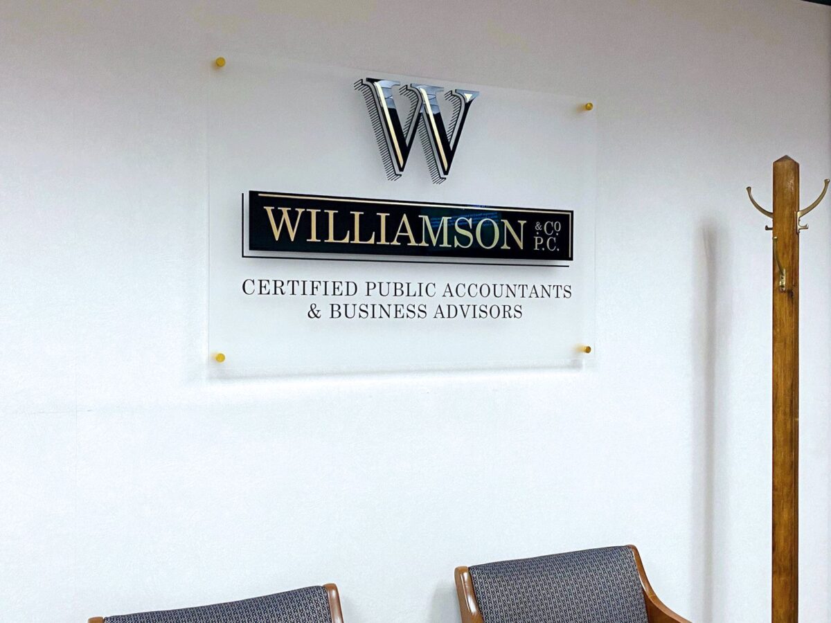 A sign on a wall reads "Williamson & Co PC, Certified Public Accountants & Business Advisors" below a metallic logo with the letter "W". Two gray chairs with wooden armrests are in front of the sign, and a wooden coat rack is on the right side.