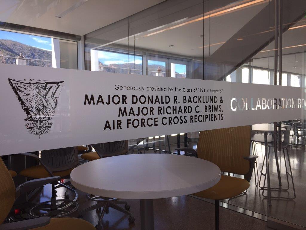 A glass door with text reading "Generously provided by The Class of 1971 in memory of MAJOR DONALD B. BACKLUND & MAJOR RICHARD C. BRIMS AIR FORCE CROSS RECIPIENTS." Several chairs and tables are visible through the glass, indicating a meeting room.