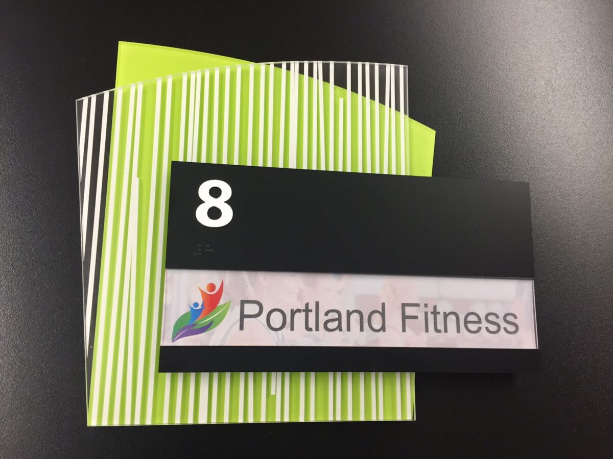 A black rectangular sign with the number "8" in white at the top. Below it, there is a colorful logo of three human figures and the text "Portland Fitness" on a white background. The sign is set against a background of vertically striped green and white paper.