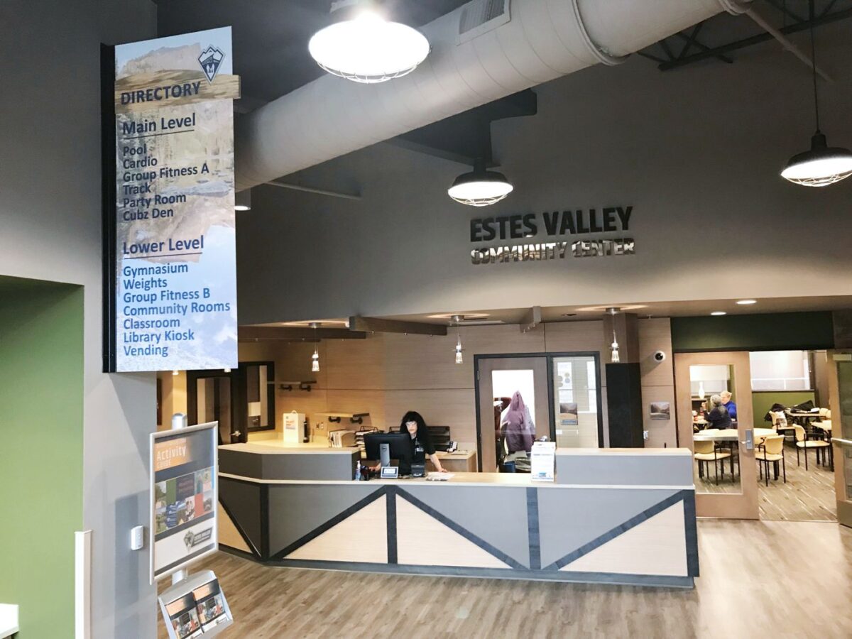 Lobby of Estes Valley Community Center featuring a reception desk with a staff member, a large directory sign listing amenities, and a prominent ceiling sign with the center's name. Background includes tables and chairs with people engaging in activities.