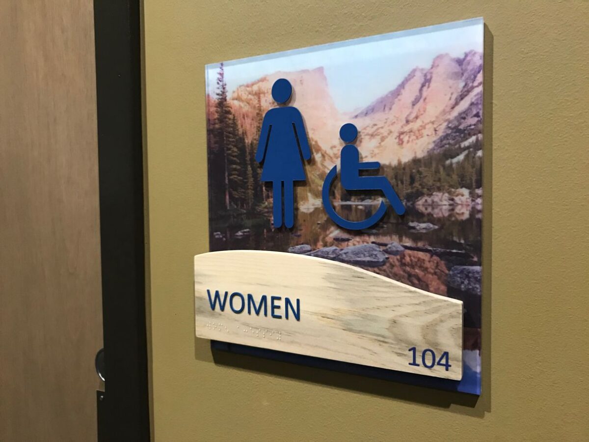 A restroom sign on a wall indicating a women's restroom, featuring symbols for women and accessibility for people with disabilities. The background includes a landscape image, and the sign is labeled "WOMEN" with the number "104" at the bottom.