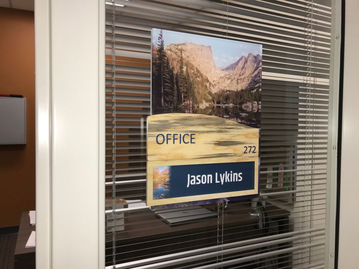 A glass door with blinds features a sign displaying a nature scene and the text "OFFICE 272 Jason Lykins." The office interior is visible through the window.
