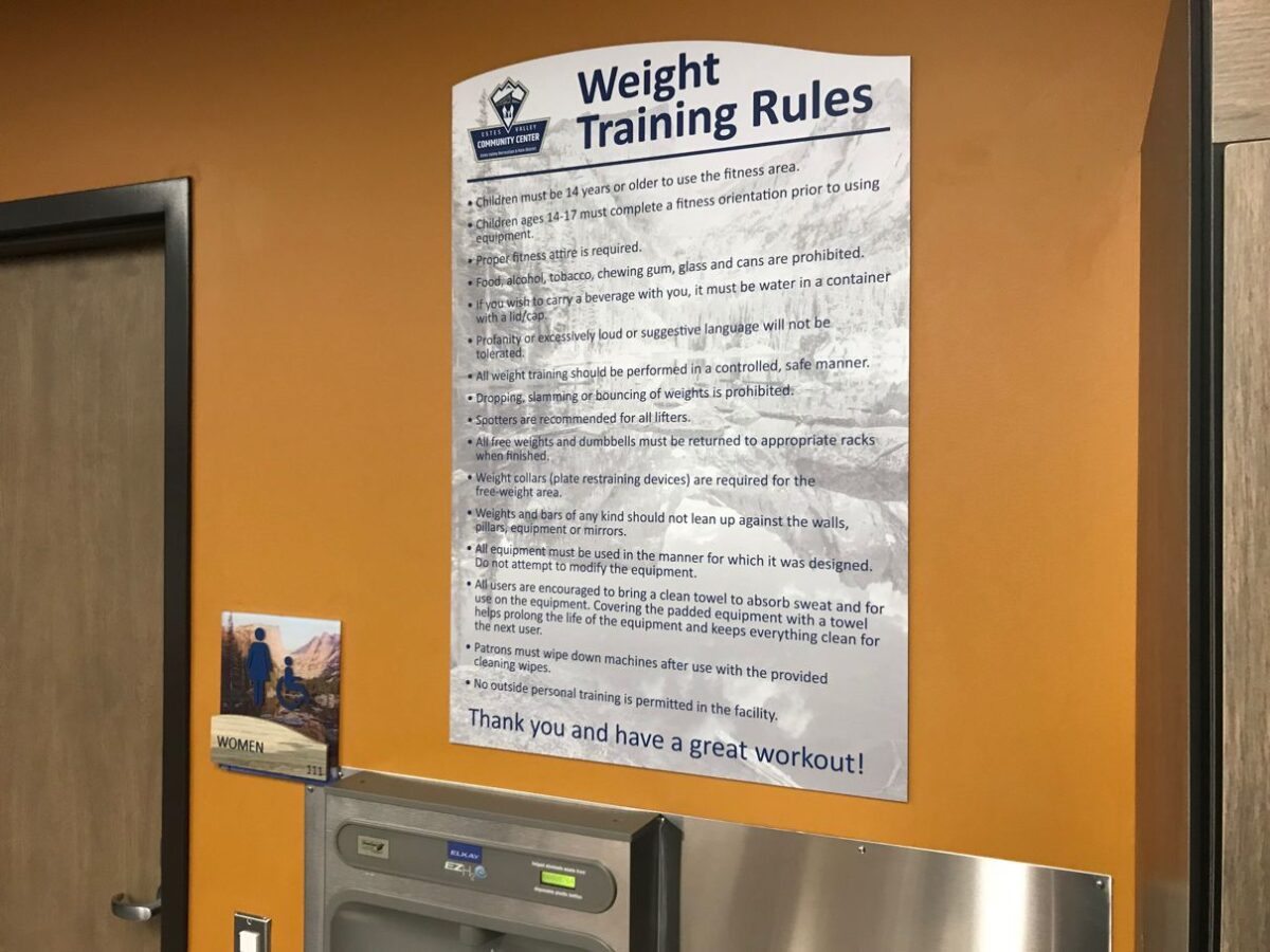 Weight training rules sign posted on an orange wall near a doorway. The sign contains various guidelines for weight training, emphasizing safety, proper equipment use, and courtesy towards other gym users. Below the sign is a water fountain and a small notice.