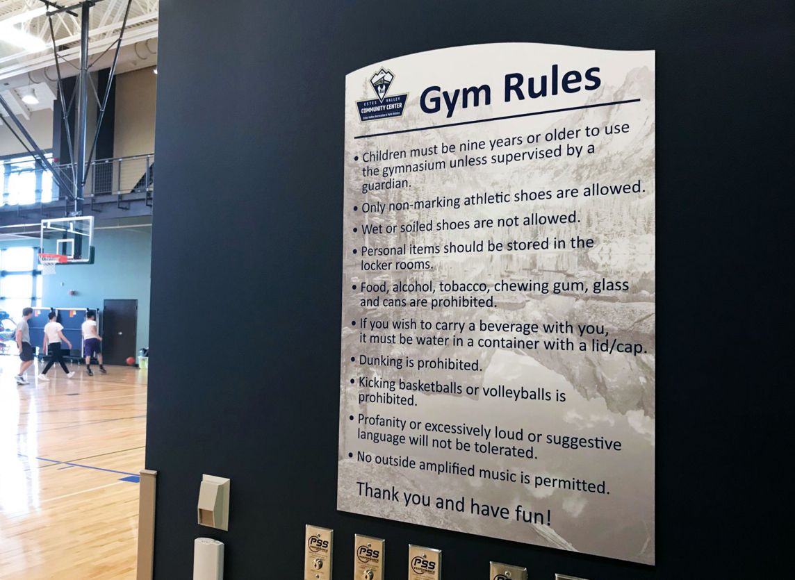 A gym rules sign is posted on a dark wall in an indoor basketball court. The rules specify not to bring gum, tobacco, food, or drinks, and highlight the requirement for appropriate attire and music. People are visible playing basketball in the background.