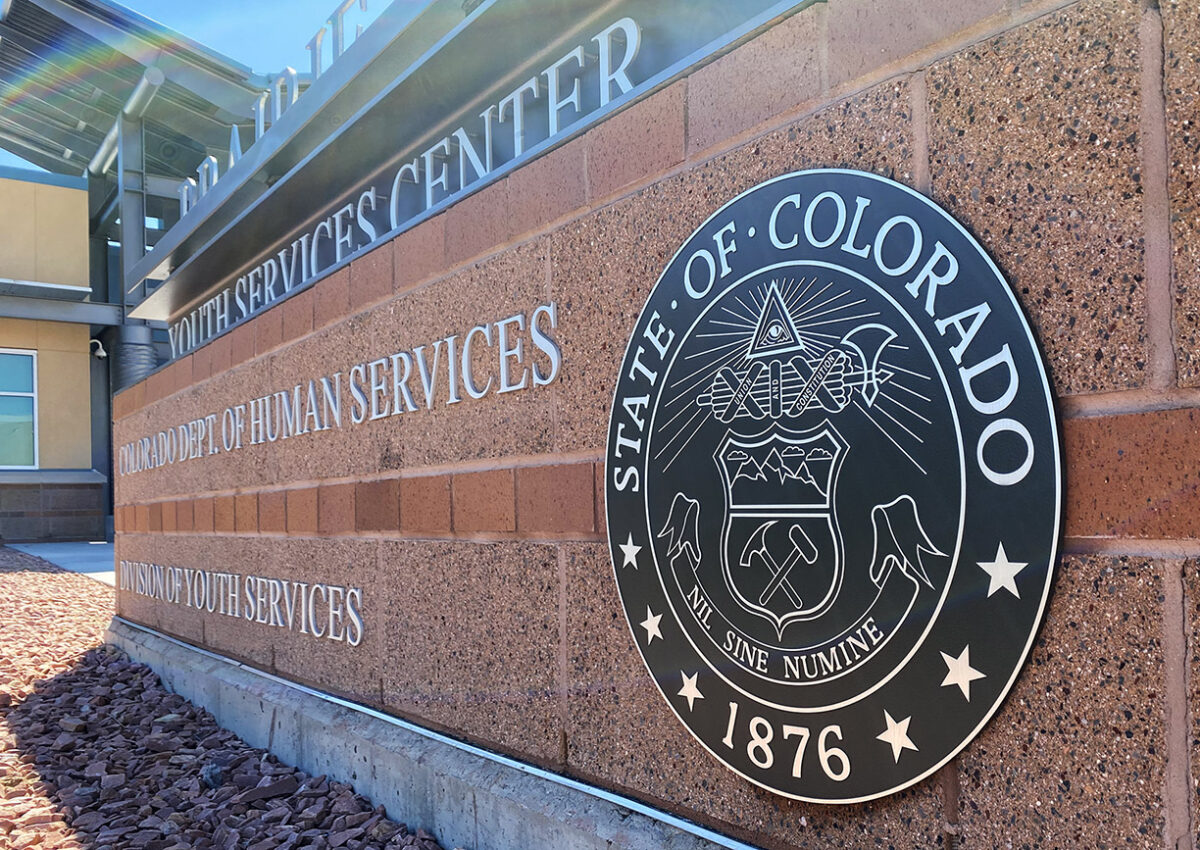A brick wall with a large circular emblem that reads "State of Colorado 1876" and the text "Department of Human Services" and "Youth Services". The emblem has various symbols, including a pickaxe, a scroll, and a Roman fasces with an axe.