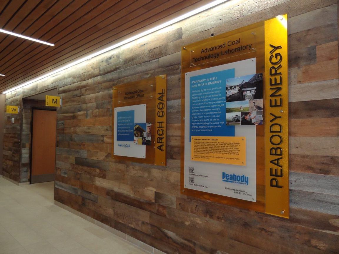 A hallway with wooden walls features two large informational posters. One poster is titled "Peabody Energy" and the other is titled "Arch Coal." Both posters contain text and images. There are doorways and directional signs also visible in the hallway.