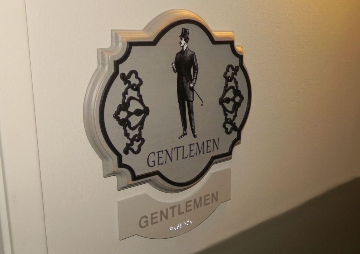 A sign on a wall indicating a gentlemen's restroom. The sign features a vintage-style illustration of a man in formal attire with a top hat and cane, and the word "GENTLEMEN" written below the illustration. The word is also repeated in braille at the bottom.