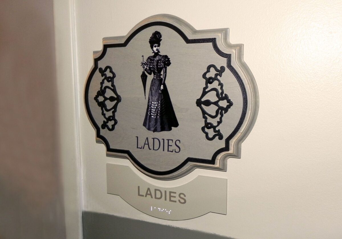 A restroom sign for ladies featuring an illustration of a Victorian-style woman in a long dress with a parasol. The sign has a decorative border and the word "LADIES" appears below the illustration.