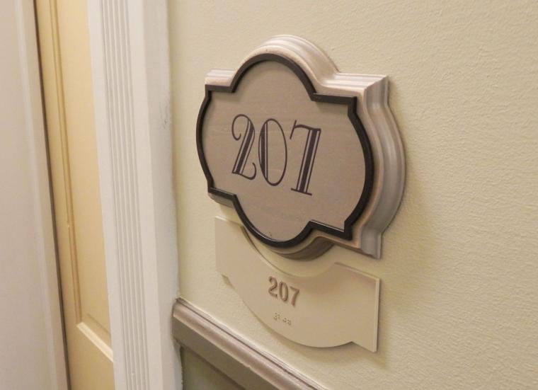 A decorative sign with the number "207" is mounted on a beige wall next to a door. Below the sign is a smaller plaque also displaying the number "207." The background includes door trim details in a light color.