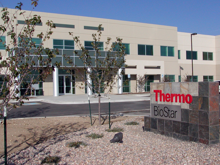 A modern, two-story commercial building with large windows and a sign in front reading "Thermo BioStar" in red and black letters. The surrounding area has young trees and a gravel landscape.