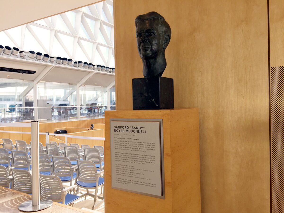 A bronze bust is displayed on a wooden pedestal next to an informational plaque. In the background, there is a bright, spacious atrium with rows of empty chairs arranged in a semicircle, large windows, and a modern architectural design.