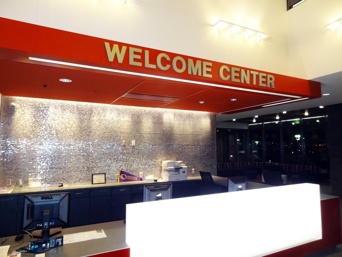 A modern welcome center with a bright red sign reading "Welcome Center" in bold white letters. The desk area below is well-lit and equipped with a computer, a printer, and other office supplies. The background features a textured metallic wall.
