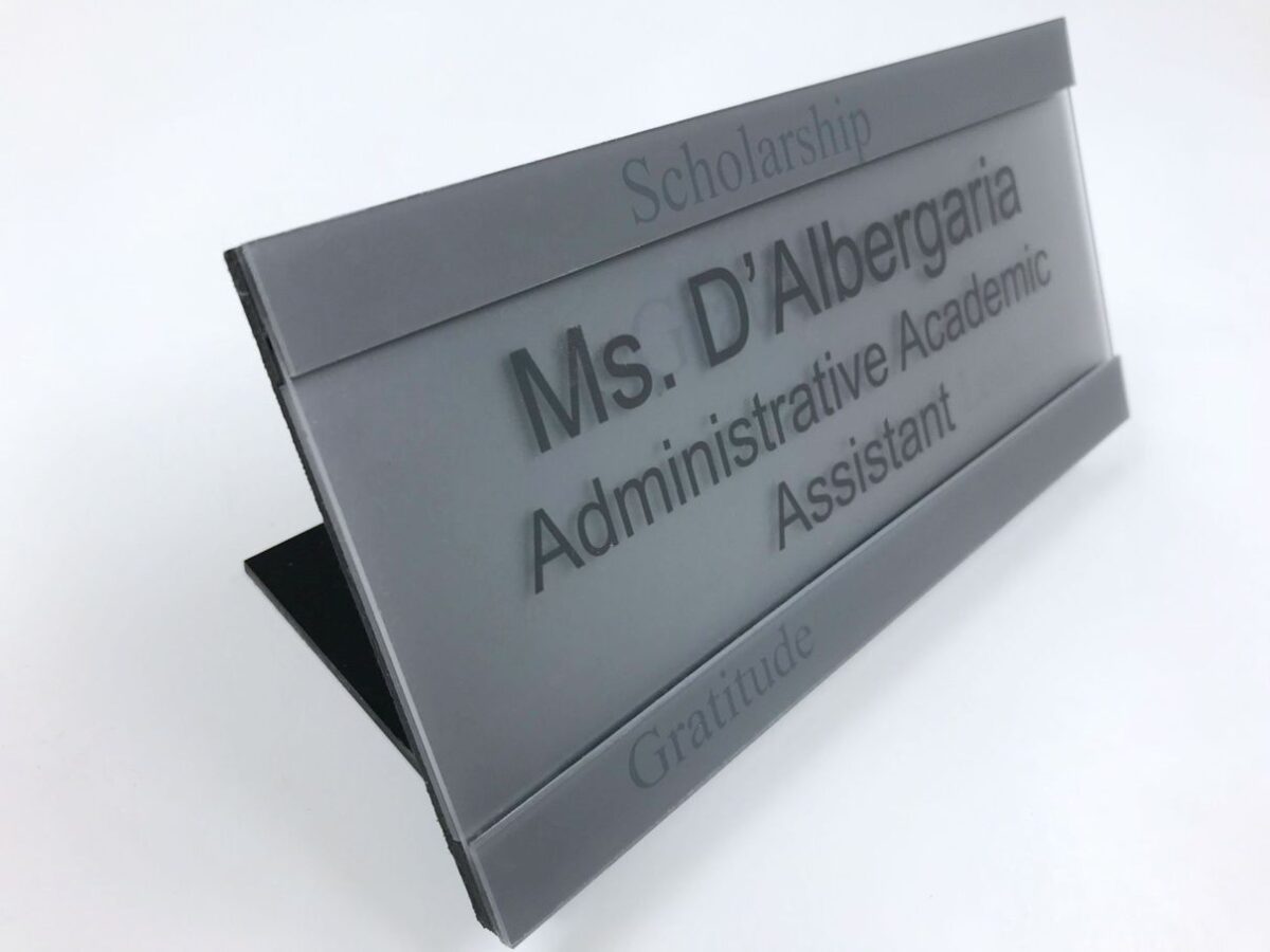 A gray desk nameplate with black lettering reads "Ms. D'Albergaria, Administrative Academic Assistant." The word "Scholarship" is engraved at the top, and "Gratitude" is engraved at the bottom.