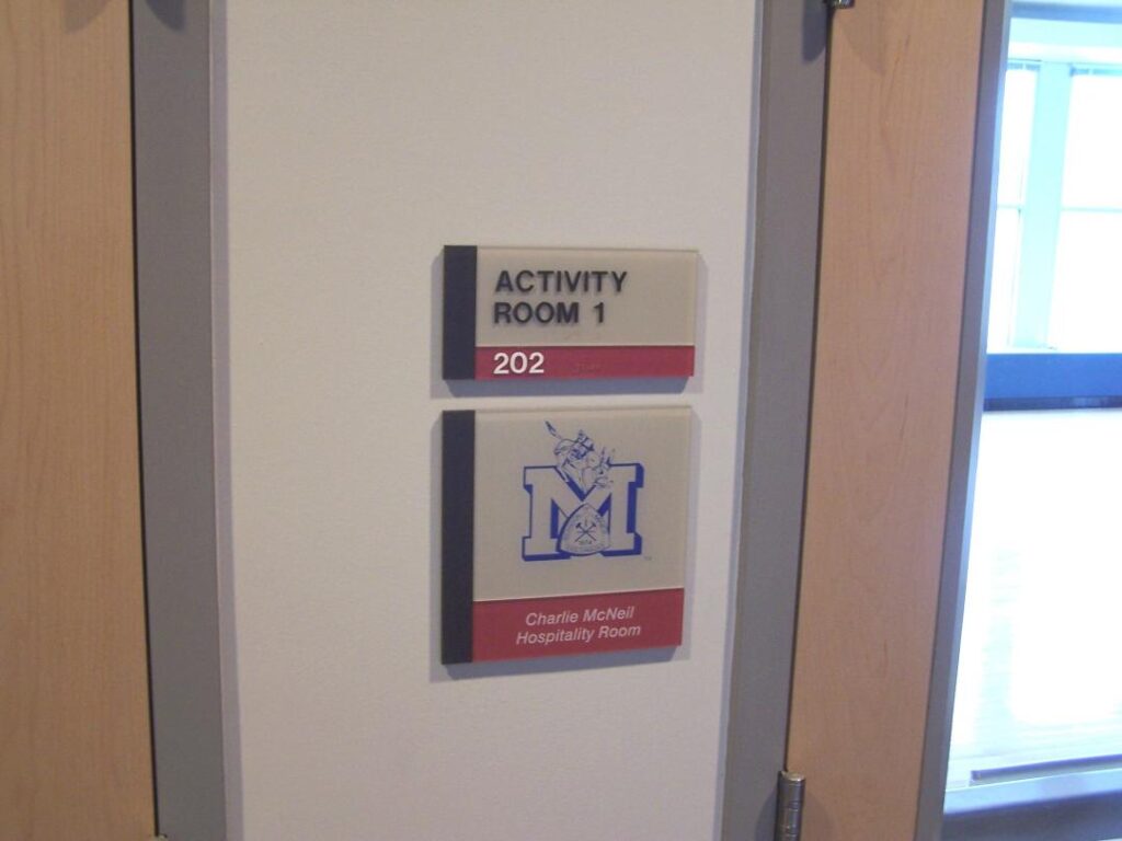 Two plaques mounted next to a door indicating 'Activity Room 1' and 'Charlie McNeill Hospitality Room,' both with room number 202 displayed.