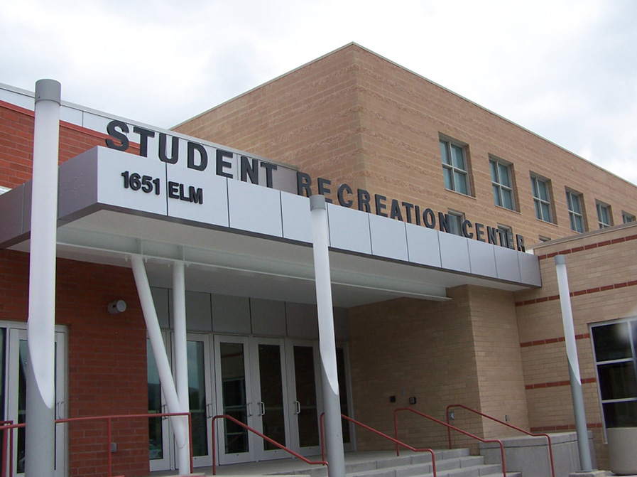 A brick building with a metal awning over an entrance reads "Student Recreation Center" with the address "1651 Elm" displayed prominently. The entrance features glass doors, and there is a red handrail along a short stairway leading to the door.