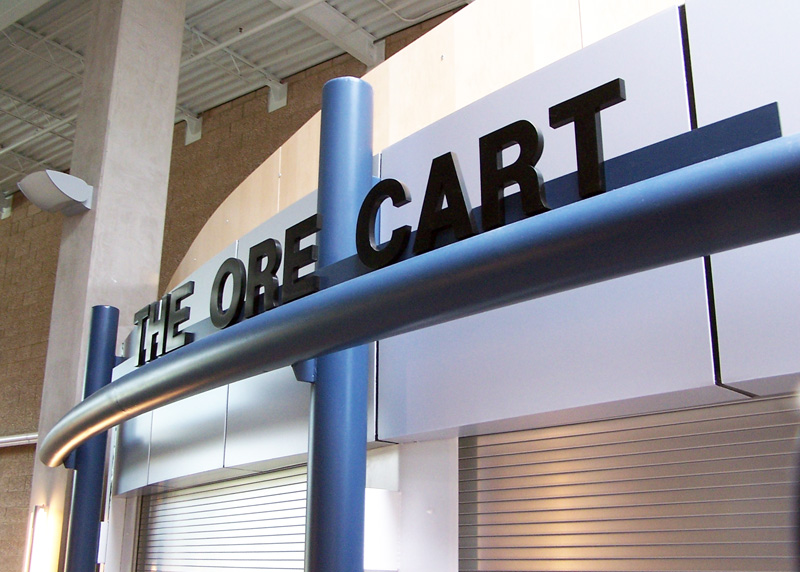A modern storefront with a sign reading "THE ORE CART." The structure features sleek blue and silver designs, with a partially visible roller shutter door.