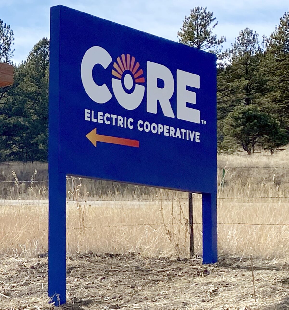 A large blue sign with the words "CORE Electric Cooperative" in white lettering and a circular logo resembling a sun. An orange arrow points to the left beneath the text. The sign is situated in a grassy area with trees in the background.