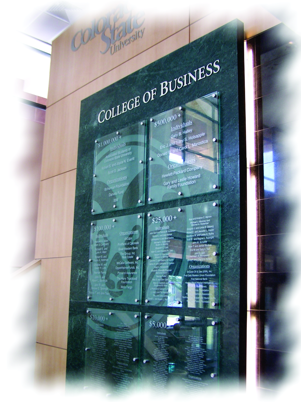 A recognition wall display at Colorado State University's College of Business listing donors and their contributions, segmented by donation amounts. The background features a mix of wood paneling and glass with etched lettering.
