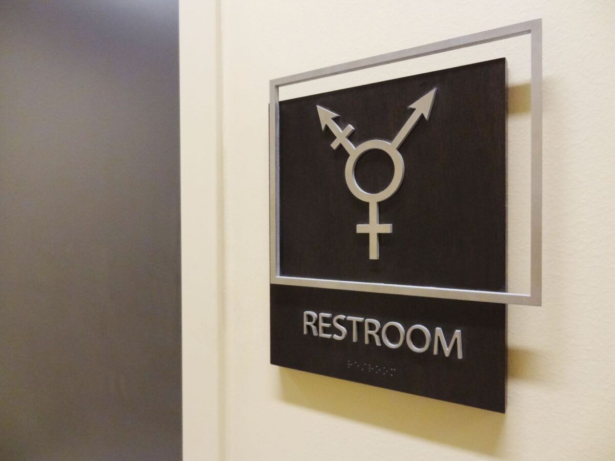 A sign on a wall denotes a restroom with a symbol combining male, female, and non-binary gender symbols. The word "Restroom" is written below the symbol.