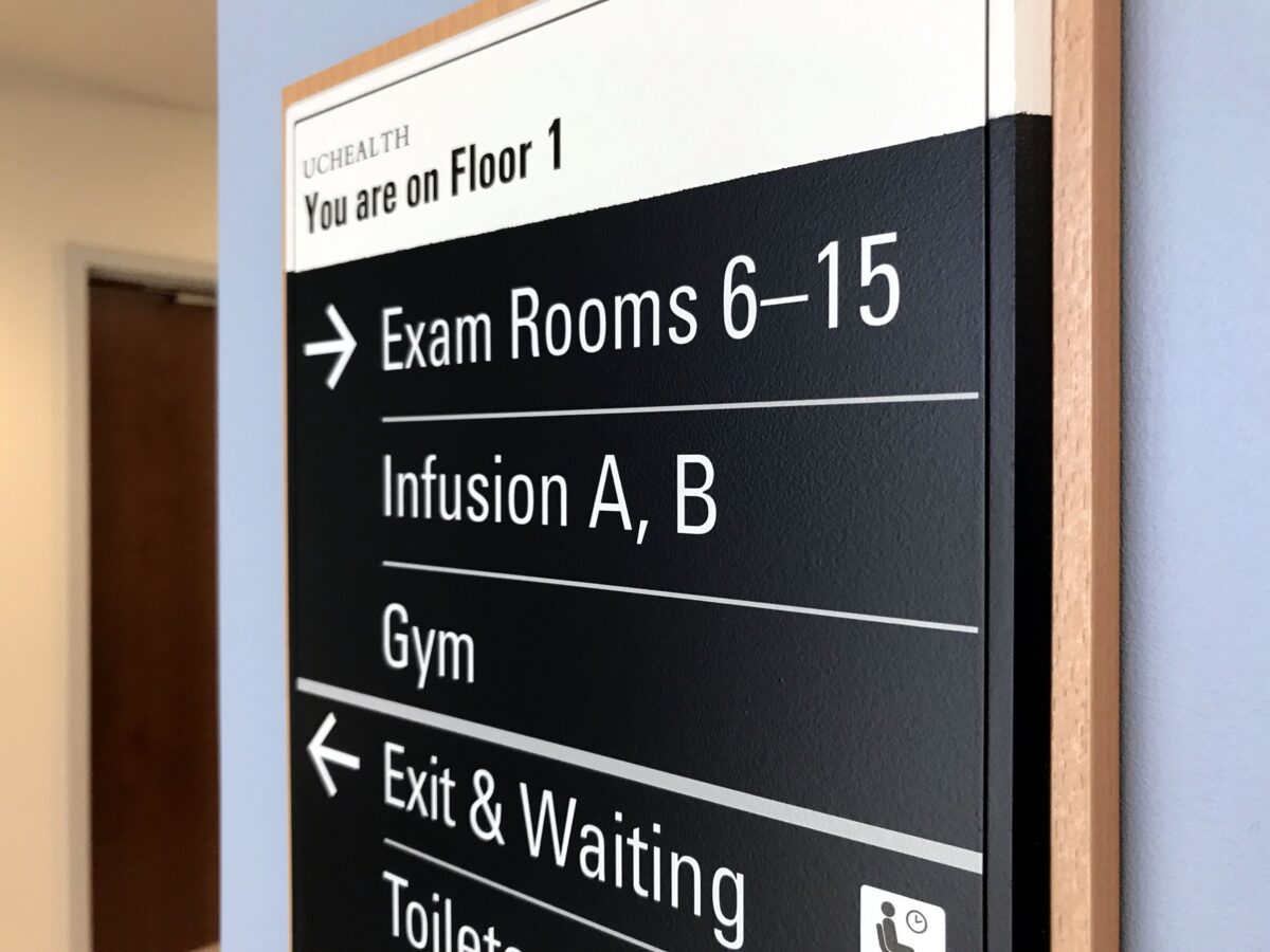 A directional sign inside a healthcare facility indicates that on Floor 1, Exam Rooms 6-15, Infusion A and B, and the Gym are to the right. The Exit & Waiting area, Toilets, and Exam Rooms 1-5 are to the left.