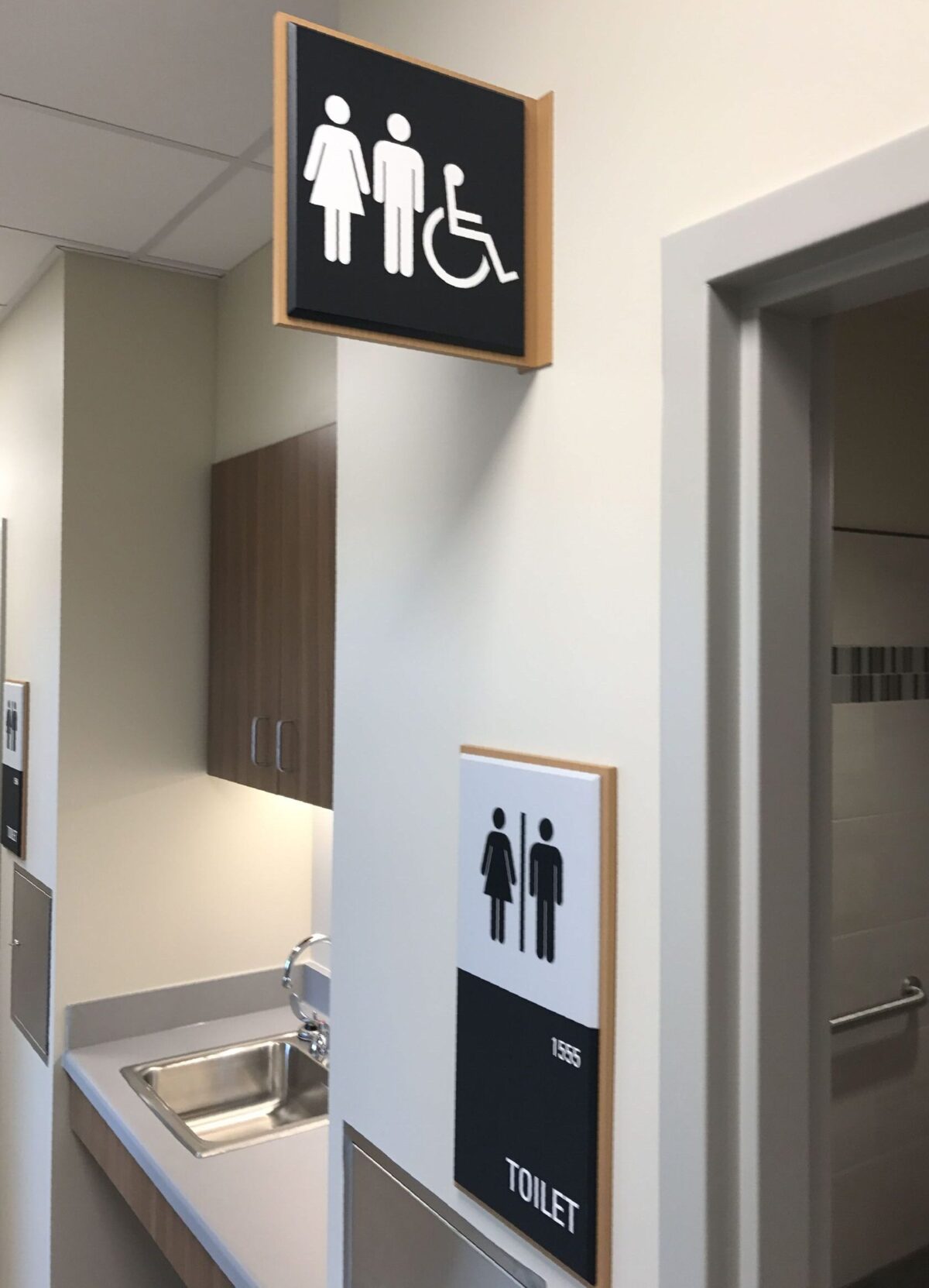 A restroom sign is mounted above a hallway, indicating a unisex toilet with accessibility features. Below it is another sign on the wall with symbols for male, female, and accessible, accompanied by the word "TOILET." A sink is visible in the background.