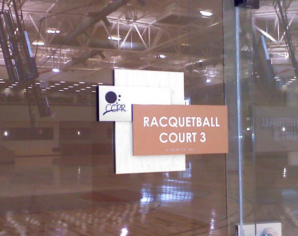 A glass door with a sign reading "Racquetball Court 3" in white text on a brown background. The interior of the racquetball court is visible through the glass door, showing the court surrounded by transparent walls and a ceiling with visible lights and beams.