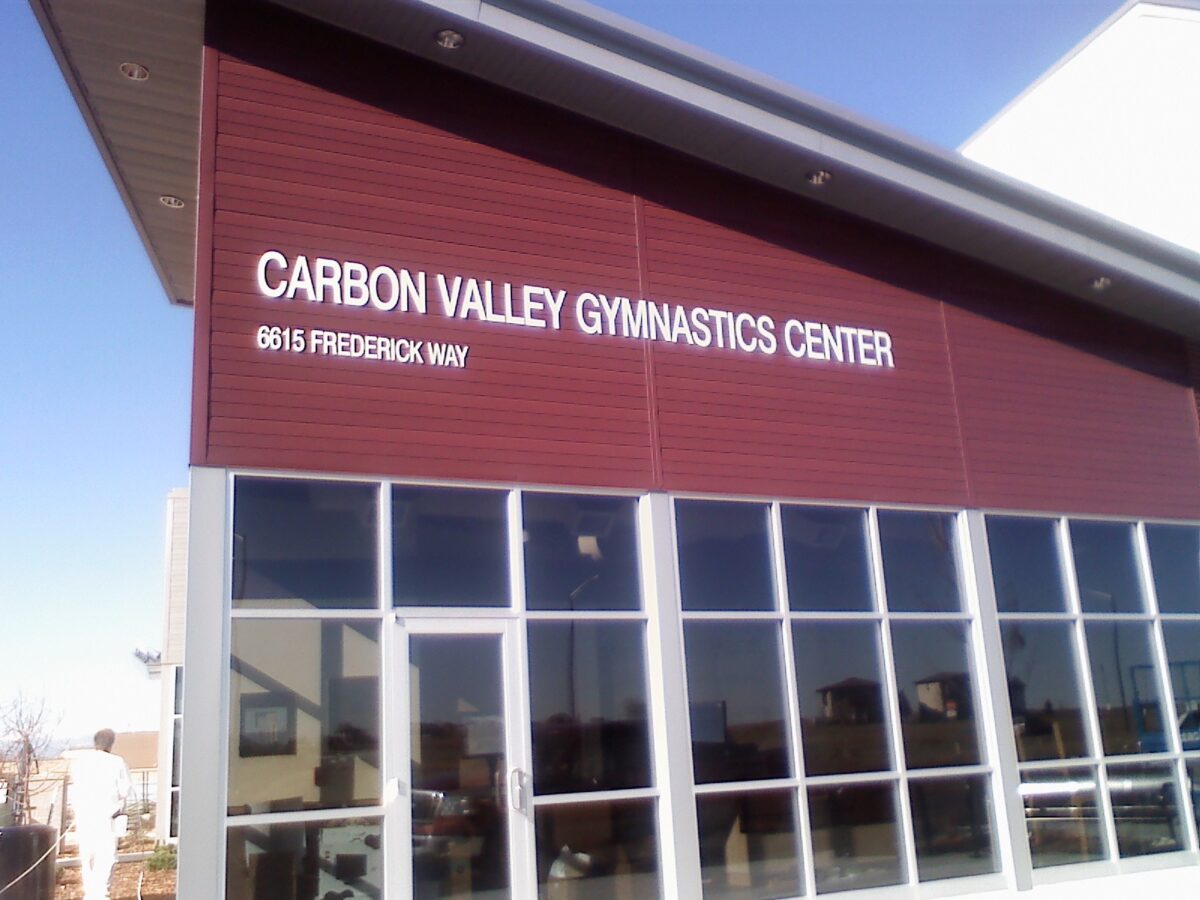 A building with a maroon-colored facade bearing the sign "Carbon Valley Gymnastics Center" in white letters. The address "0615 Frederick Way" is also displayed below the sign. The lower half of the building features large glass windows.