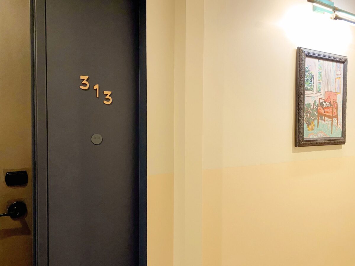 A dark gray door with the number "313" is located on the left side of a beige hallway. To the right of the door, a framed painting depicting a room with a red chair and a table hangs on the wall. The hallway is illuminated by overhead lighting.