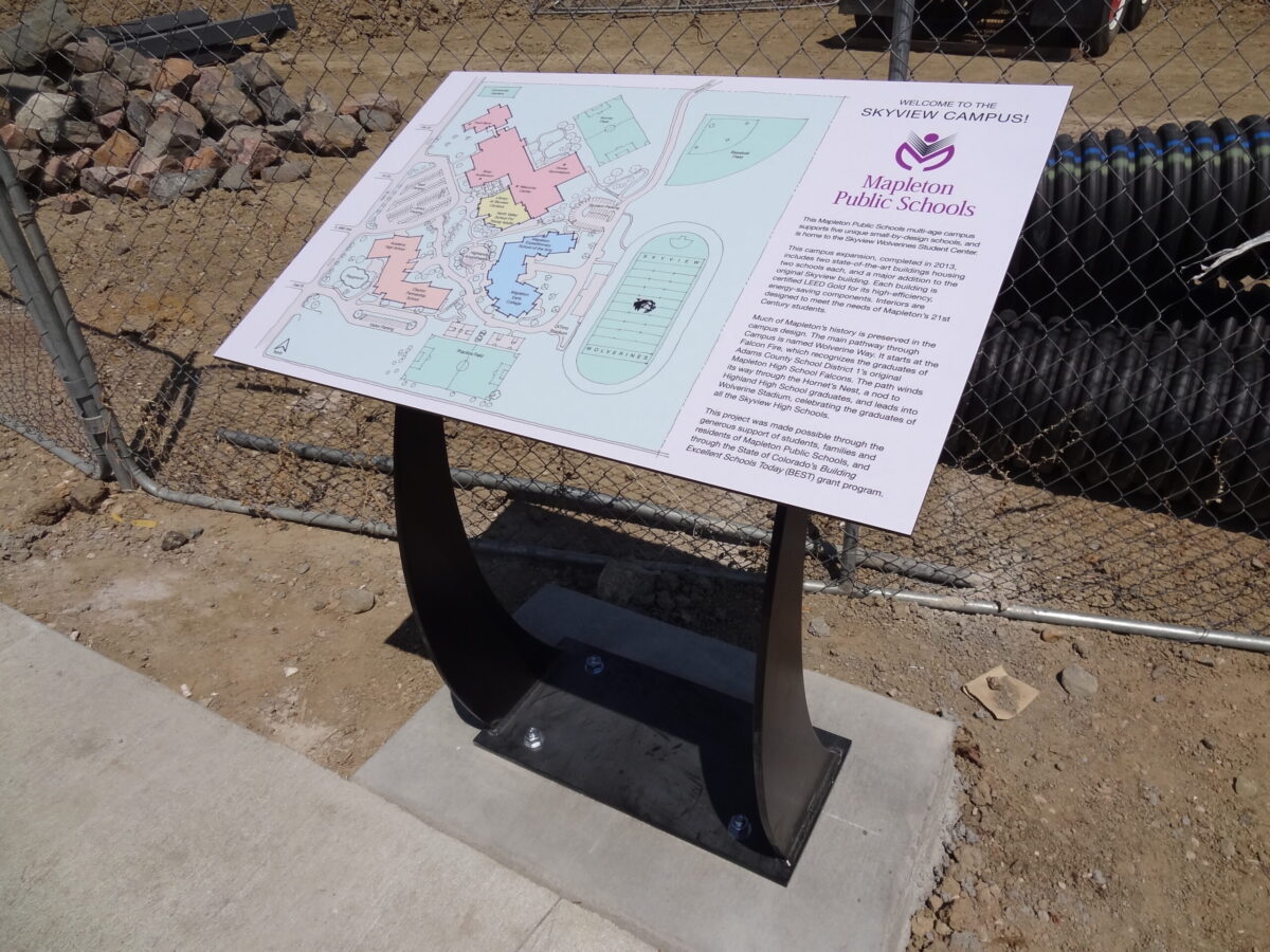 An outdoor informational sign and map on a metal stand for Alpine Public Schools, placed on a concrete slab beside a chain-link fence and construction materials. The sign contains colorful sections and text for visitors to read.