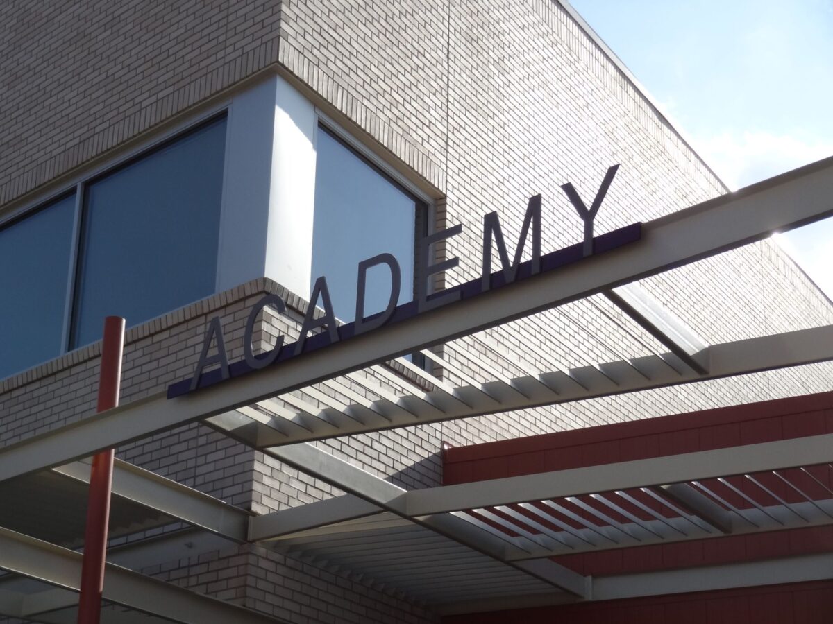 A modern building with large windows has an overhang featuring a sign with the word "ACADEMY" in capital letters. The structure is made of brick and metal, and the sky is clear in the background.