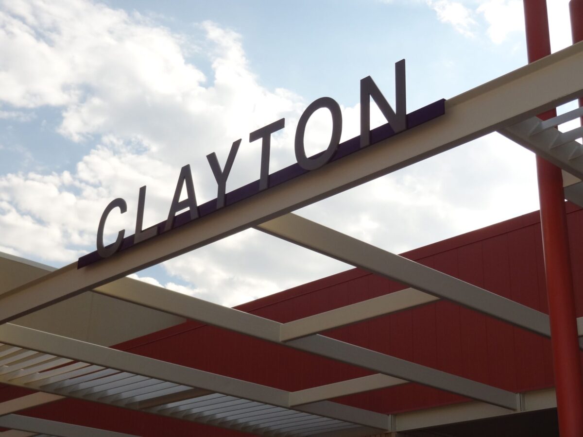 A metal structure with "CLAYTON" displayed in large, capital letters against a partially cloudy sky. The building features a mix of red and beige colors, and the photograph is taken from an upward angle.