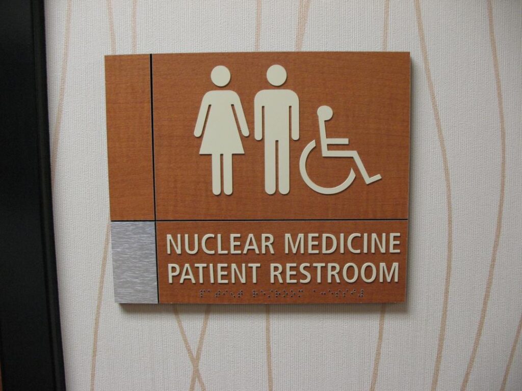 Restroom sign for 'Nuclear Medicine Patient Restroom' featuring gender symbols and a wheelchair accessibility icon on a wooden plaque.