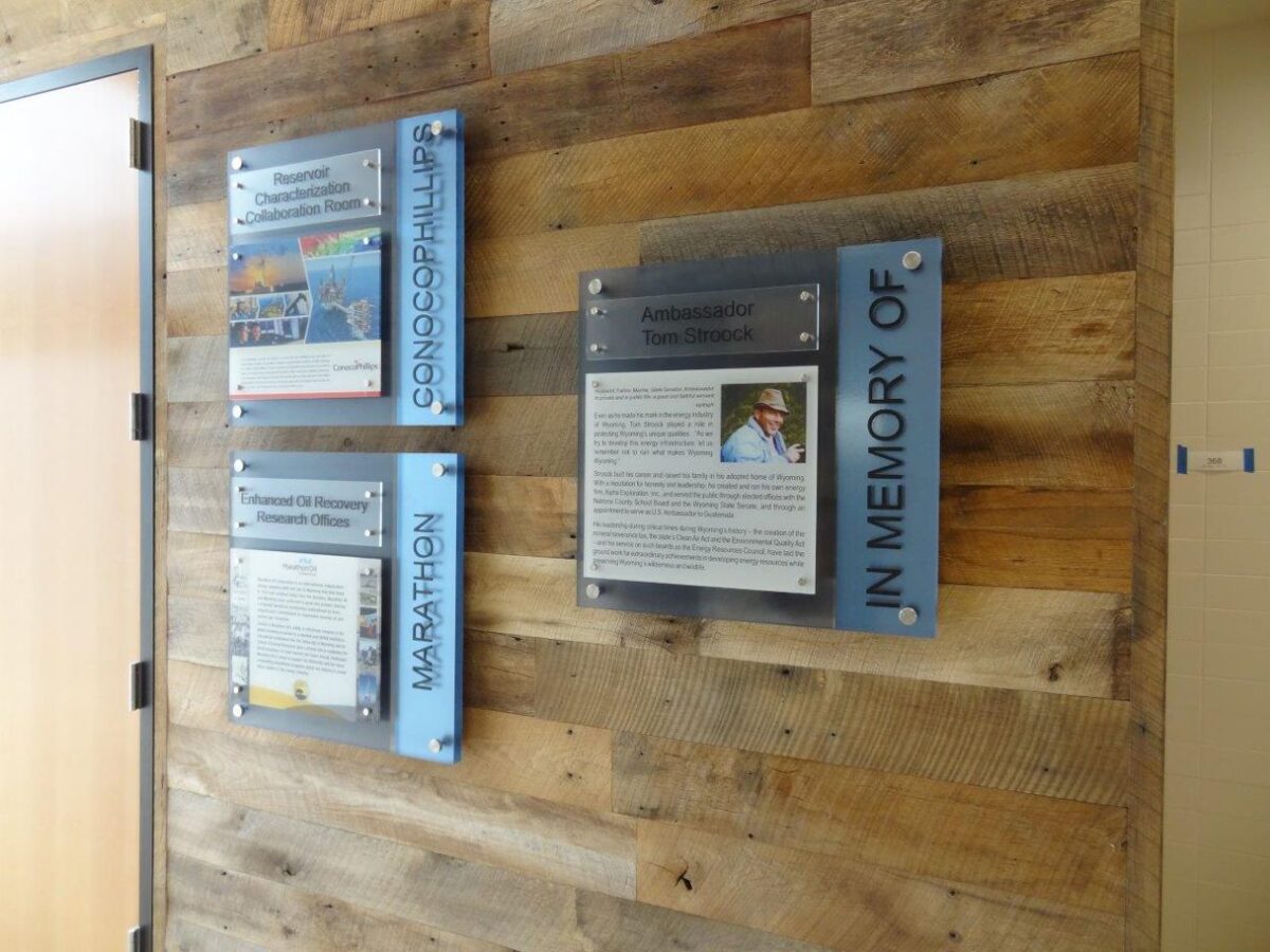 Three plaques are mounted on a wooden wall. They contain text and photos commemorating various subjects. One plaque includes the phrase "IN MEMORY OF." Each plaque is framed with transparent and colored borders, enhancing their prominence on the rustic surface.