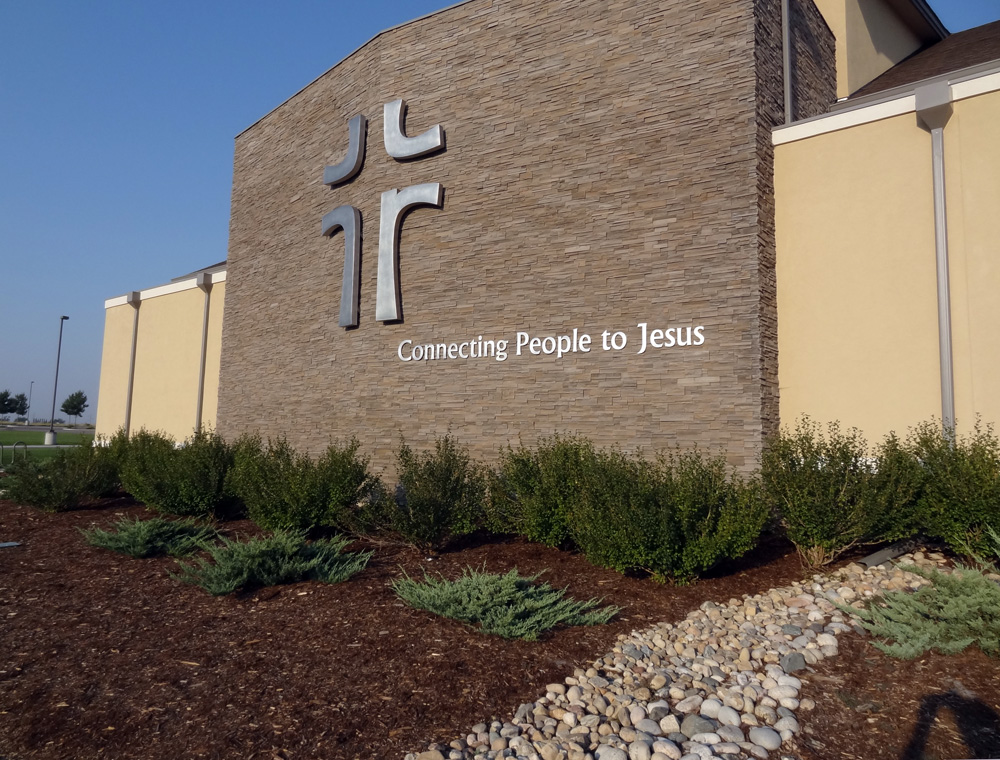 A church building with a stone facade and an emblem resembling crosses on the upper left. Below the emblem, the text "Connecting People to Jesus" is displayed. There are well-maintained bushes and a gravel path in front of the building.