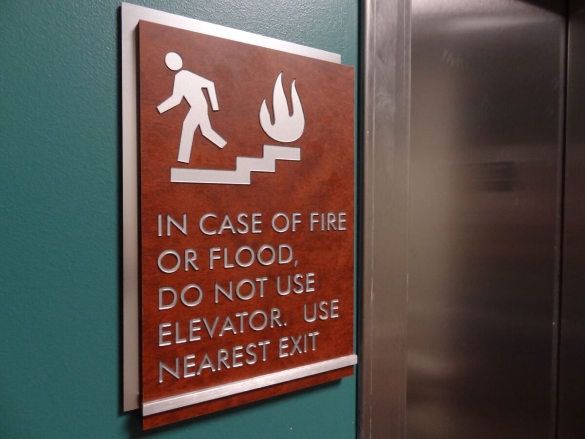 A wall sign next to an elevator depicts a person descending stairs with a flame icon. The text reads: "In case of fire or flood, do not use elevator. Use nearest exit." The sign has a brown background and white text and graphics.