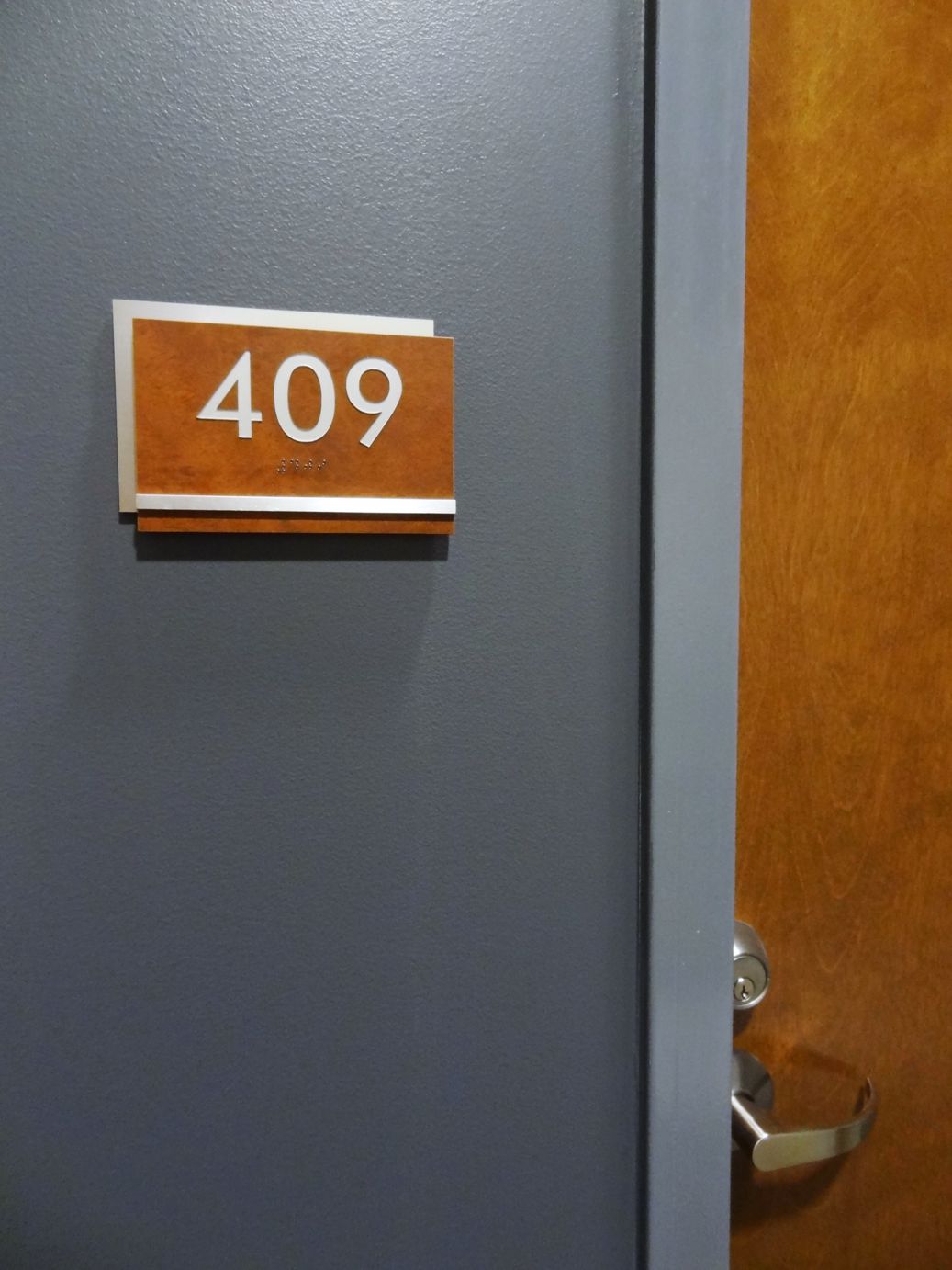 A grey door with a room number sign that reads "409" in white text on a rust-colored background is partially open, revealing part of a wooden door frame and a metal door handle.