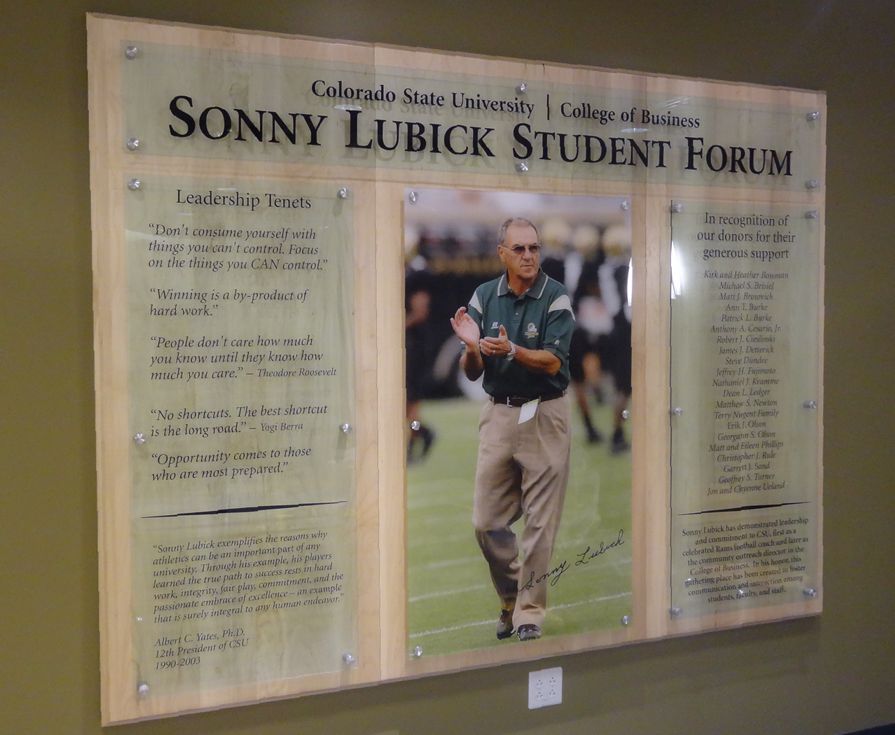 A wall-mounted wooden plaque titled "Sonny Lubick Student Forum" at Colorado State University College of Business, featuring an image of a man in the center with lists of leadership traits on both sides. The plaque highlights leadership qualities and principles.