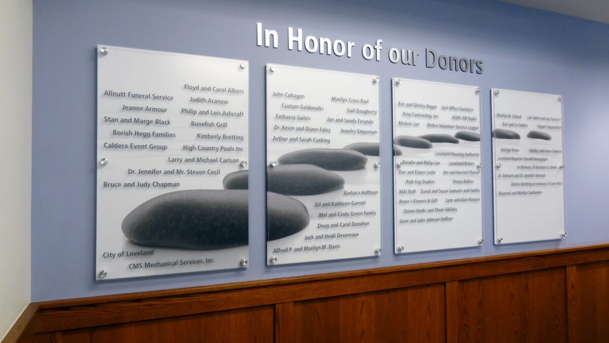 Four plaques on a wall honor donors. Each plaque lists multiple names under the heading "In Honor of Our Donors." The plaques are mounted on a light blue wall above a wooden panel.