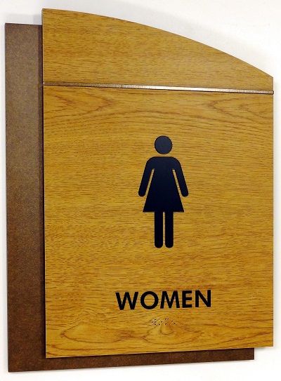 Wooden restroom sign indicating a women's bathroom. The sign has a simple silhouette of a woman and the word "WOMEN" written below in black text. The sign has a rectangular shape with a slightly slanted top.