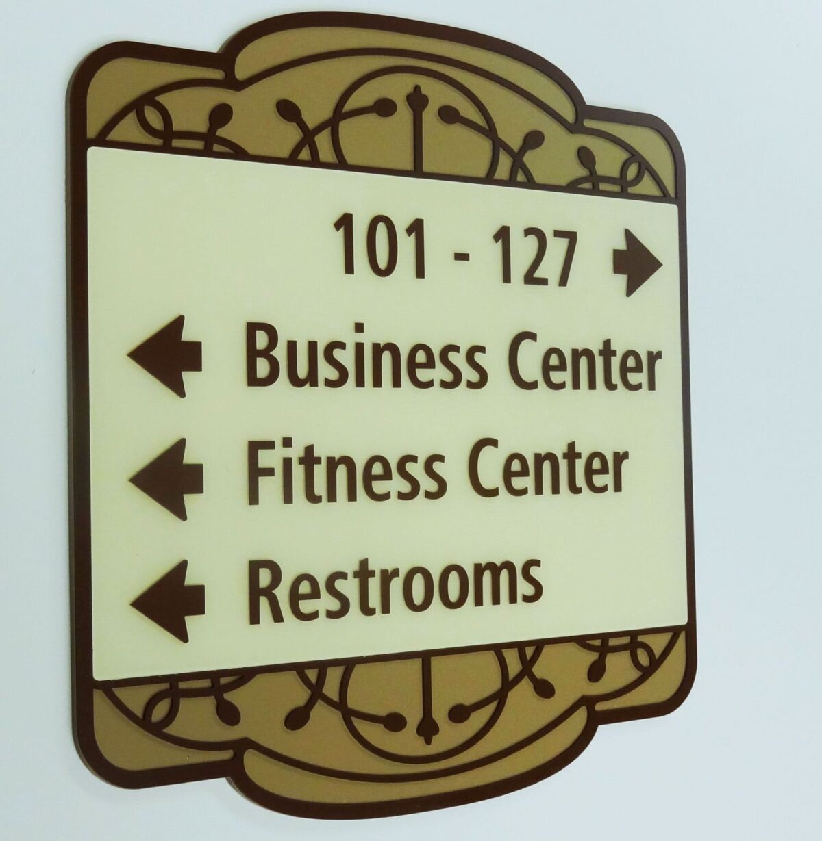A wall-mounted directional sign with an ornate design points arrows in various directions. To the left are directions to the Business Center, Fitness Center, and Restrooms. An arrow pointing right leads to rooms numbered 101 - 127.
