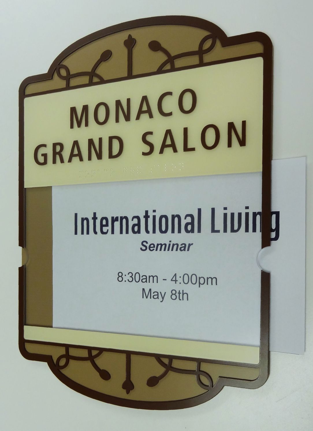 A sign for the "Monaco Grand Salon" with an insert underneath that reads, "International Living Seminar, 8:30am - 4:00pm, May 8th." The sign has a decorative border and the text is in a mixture of brown and black fonts on a beige background.