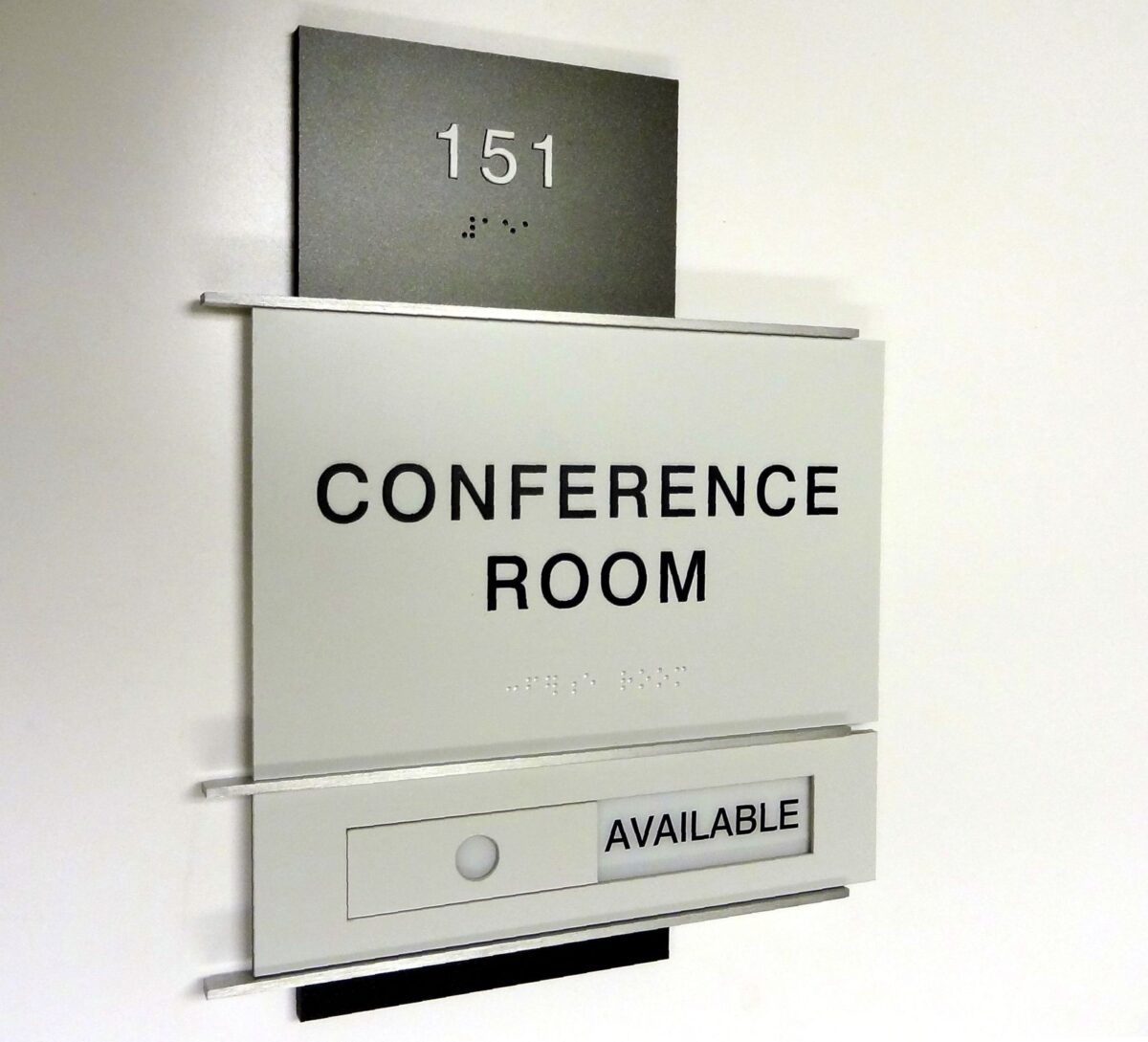 A white and grey conference room sign with the number 151 and Braille text at the top. The middle section reads "CONFERENCE ROOM," and a small sign at the bottom indicates "AVAILABLE" with a green dot next to it.