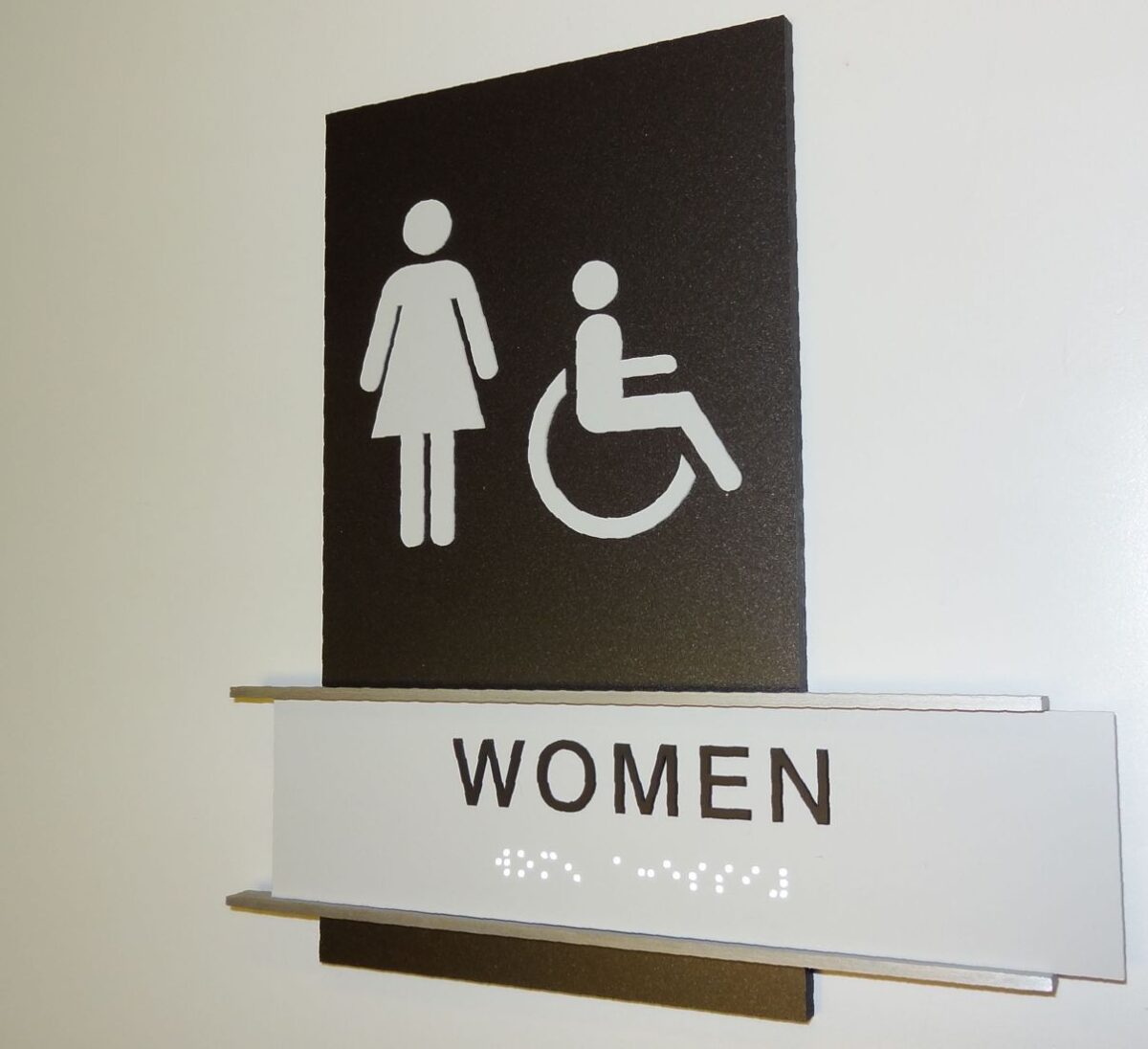 A restroom sign on a wall indicating facilities for women, including those with disabilities. The sign has symbols for a standing woman and a person in a wheelchair above the word "WOMEN". Below the text, there is Braille for accessibility.