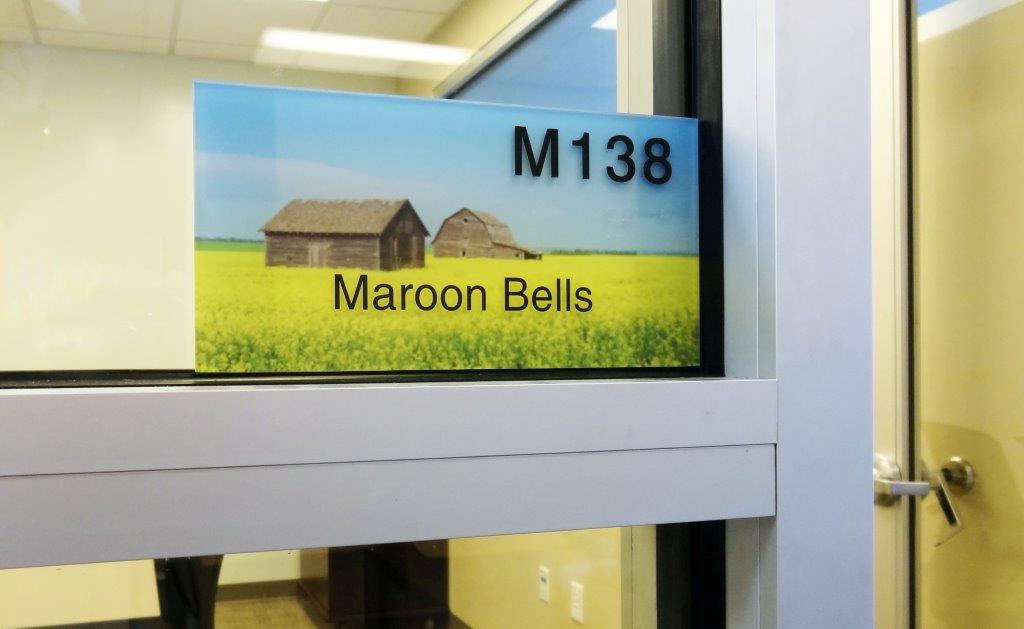A room nameplate with the designation "M138" and the title "Maroon Bells." The sign includes an image of two barns in a field with yellow flowers, set against a backdrop of blue sky. The nameplate is affixed to a glass door with a white frame.