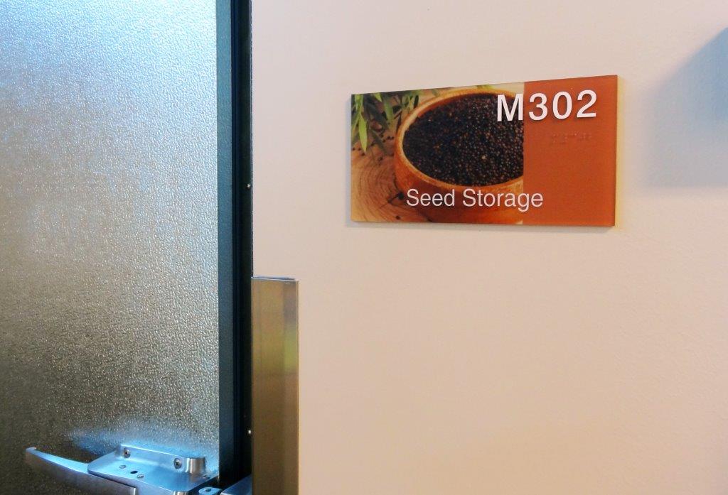 A door with a frosted glass panel is partially open. A sign next to the door reads "M302 Seed Storage" and features an image of a bowl filled with black seeds.