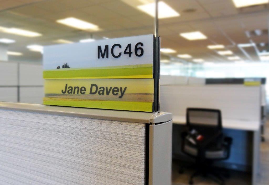 An office cubicle is labeled with a nameplate reading "MC46" and "Jane Davey." The cubicle is empty, featuring a chair and desk inside. The surrounding area includes similar cubicles under fluorescent lighting.